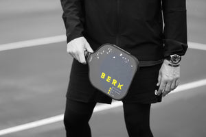 Berk Athlete holding The Calafia paddle in the backhand position standing infront of the pickleball net. The Calafia has the Berk logo in yellow with some splattered paint on the opposite side. The rest of the paddle is black. Designed for Power. 