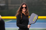 Pretty Brunette in black jacket and oversized sunglasses holding the Berk Calafia paddle in the black colorway. She has a big smile on her face. The Calafia is a premium pickleball paddle with a solid core designed for power and control. 