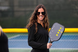 Pretty Brunette in black jacket and oversized sunglasses holding the Berk Calafia paddle in the black colorway. She has a big smile on her face. The Calafia is a premium pickleball paddle with a solid core designed for power and control. 