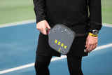 Berk pickleball Athlete stands holding the Berk Calafia paddle infront of waist. Athlete is in all black holding the Berk pickleball Calafia paddle in the black colorway. He wears a diver's watch on his left wrist. 