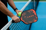 Berk athlete standing with hands on the pickleball net holding the Berk Calafia premium pickleball paddle in Fire colorway in his right hand. He is holding a pickleball in his left hand. 