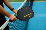 Athlete standing with hands on the net holding the Berk Calafia paddle in the black colorway. In left hand he holds a pickleball. The Calafia is a solid core pickleball paddle designed for power and large sweet spot that wont breakdown over time. 
