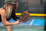 Pretty Blond girl holding the Berk Calafia fire paddle getting ready to hit the ball. She is wearing an army green hat, sunglasses and smiling. The Calafia paddle has a rough textured graphite face, a low profile edge guard for increased protection and a premium textured grip for increased traction and elevated moisture wicking. 